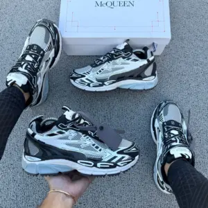 Zapatillas urbanas Off-White blancas y negras con diseño técnico contemporáneo, suela gruesa y detalles reflectantes, mostradas en pie y mano sobre superficie gris.